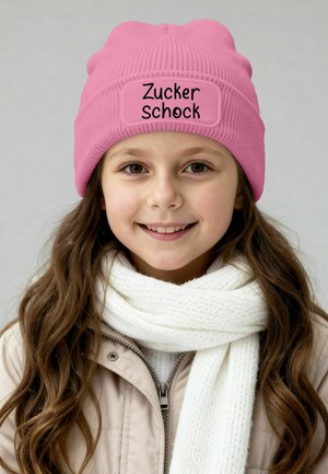 Pink knit beanie with a rectangular patch labeled "Zucker Schock." Paired with a white, textured scarf and a light beige jacket.