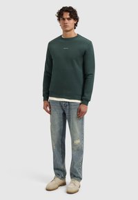 Groene sweatshirt met ronde hals en geribbelde boorden, gedragen over een wit shirt. In combinatie met lichtblauwe distressed jeans en off-white schoenen.