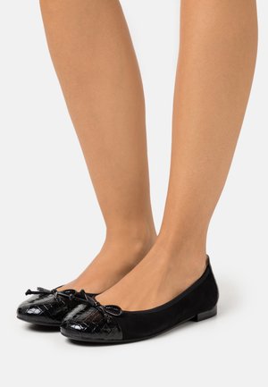 Ballerines noires en simili cuir verni texturé avec des bouts ornés de petits nœuds, portées pieds nus sur fond blanc.