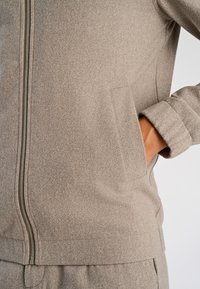 Jasnobrązowa kurtka na zamek wykonana z teksturowanego materiału, z bocznymi kieszeniami i podwiniętymi rękawami. Szczegółowe przeszycia wokół zamka.