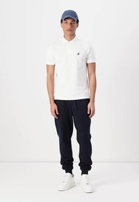 Camiseta polo blanca con un logotipo, combinada con pantalones jogger azul marino. El modelo lleva una gorra azul y zapatillas blancas, de pie sobre un fondo sencillo.