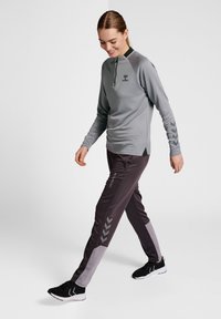 Maglia grigia chiara a maniche lunghe con colletto a zip, abbinata a pantaloni viola scuro con un pannello grigio chiaro e un motivo a chevron sul lato.