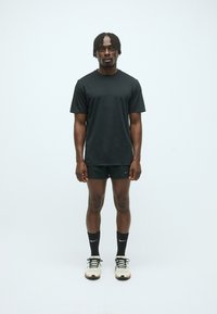 T-shirt noir à manches courtes et short noir, tous deux en tissu lisse. Chaussures de sport avec accents blancs et chaussettes noires. Fond neutre.