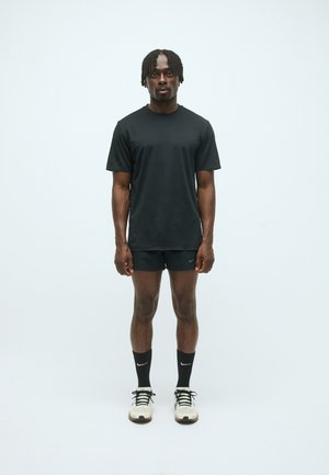 Schwarzes kurzärmeliges T-Shirt und schwarze Shorts, beide aus glattem Stoff. Sportliche Schuhe mit weißen Akzenten und schwarzen Socken. Neutraler Hintergrund.