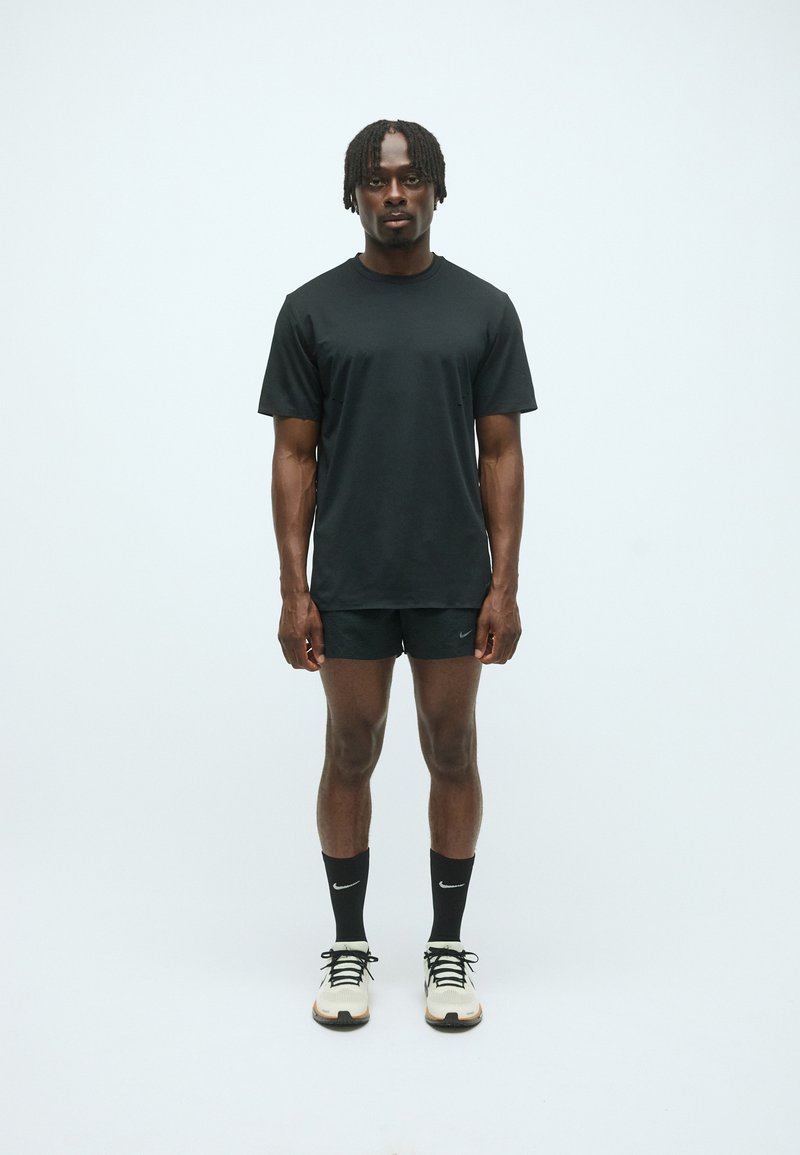 T-shirt noir à manches courtes et short noir, tous deux en tissu lisse. Chaussures de sport avec accents blancs et chaussettes noires. Fond neutre.