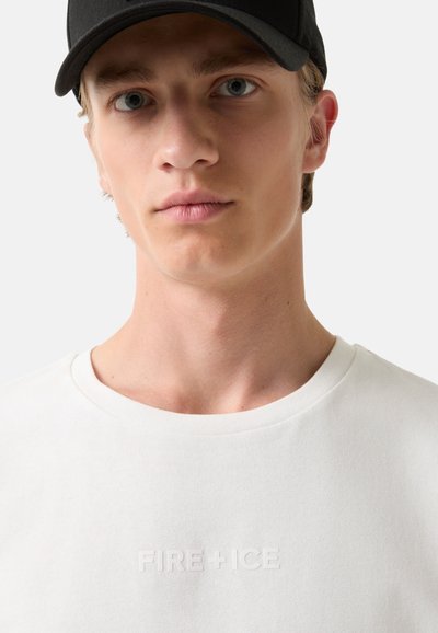 Bílý krátký rukáv tričko s kulatým výstřihem a decentně embosovaným logem "FIRE+ICE" na hrudi. Model nosí černou kšiltovku.