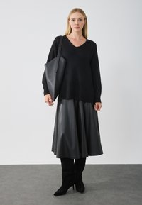 Donna che indossa un maglione nero a scollo a v, una gonna midi in pelle nera, stivali neri e porta una grande borsa a spalla nera su uno sfondo grigio uniforme.