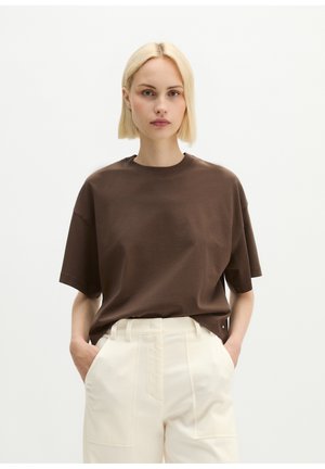 Jonge vrouw met kort blond haar, een los bruin t-shirt gestoken in een hooggetailleerde crème broek met handen in de zakken.