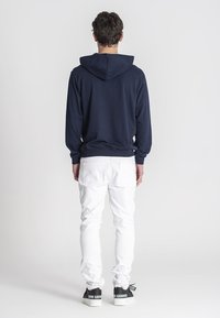 Námornícka mikina s kapucňou na sťahovanie, rebrovanými manžetami a spodným okrajom, skombinovaná s bielymi džínsami a čiernymi teniskami s logom na zadnej strane.