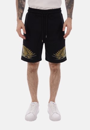 Uomo che indossa pantaloncini neri con disegni a ali gialle e verdi sui lati, una maglietta bianca e scarpe da ginnastica bianche, in piedi con le braccia lungo i fianchi.