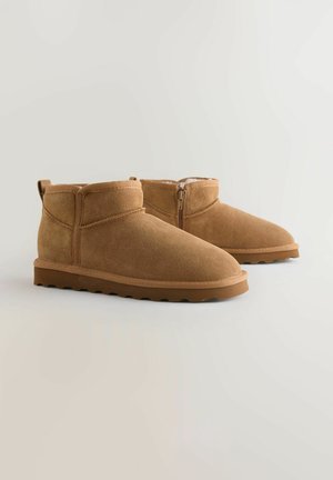 Bottines en suède beige avec une finition lisse, bout rond et fermeture éclair sur le côté. La semelle présente un design texturé et bas.