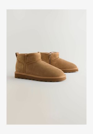 Botas de tobillo de ante en tono marrón claro con acabado suave, punta redondeada y cierre de cremallera en el lateral. La suela presenta un diseño bajo y texturizado.
