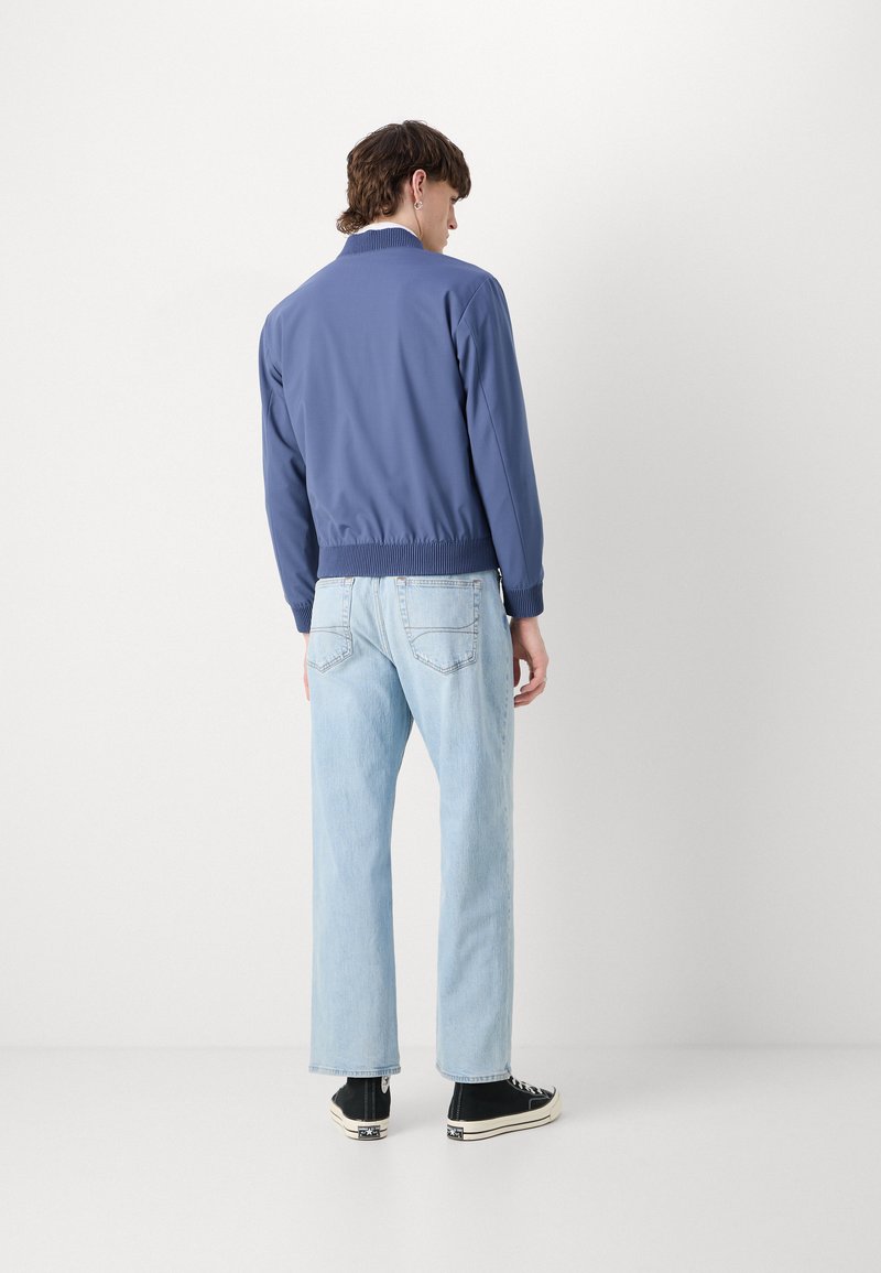 Veste légère bleue avec un col et des poignets côtelés, associée à un jean en denim bleu clair et à des baskets montantes noires.