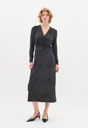 Svart långärmad klänning med djup V-ringning, prydd med texturerade vågmönster och en sidslits. Matchad med svarta spetsiga klackar.