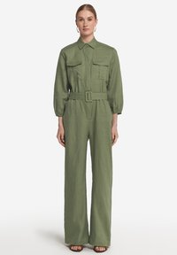 Olivgrön linne jumpsuit med långa ärmar, knappstängd krage, två bröstfickor och midja med bälte. Design med vida ben.