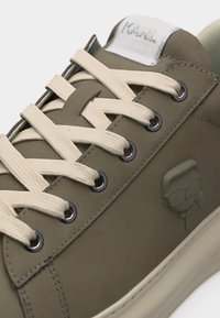 Αθλητικό παπούτσι σε ελιά πράσινο με κορδόνια σε κρεμ απόχρωση, μεταλλικά οφθαλμάκια, ανάγλυφο σήμα με στυλιζαρισμένο πρόσωπο στο πλάι και ετικέτα με το λογότυπο στη γλώσσα.