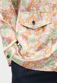 Τσέπη από μπουφάν με λουλουδένιο σχέδιο σε πορτοκαλί, κρεμ, λιλά και μέντα, με μαύρο κουμπί, φορεμένο πάνω από σκούρο παντελόνι με ριγέ λεπτομέρειες.