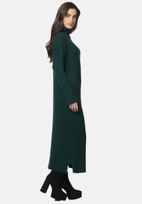 Donkergroene ribgebreide jurk met een hoge hals, splitten aan de zijkant en lange mouwen, gedragen met zwarte enkellaarsjes met hoge hak.