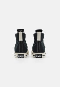 Zapatillas altas, material de lona negra, suelas de goma blanca, costuras en contraste, etiqueta con la marca en el talón y ojales metálicos para los cordones.