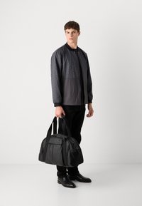 Black bomber jacket with hexagonal pattern, paired with black trousers and shoes. Holding a black duffel bag with embossed logo.