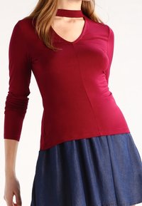 Atasan lengan panjang burgundy dengan leher V dan detail choker, memiliki desain yang fitted dan tekstur yang halus. Dipadukan dengan rok denim.