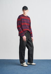 Μπλούζα με ριγέ μακριά μανίκια σε κόκκινο και σκούρο μπλε, σε συνδυασμό με άνετο μαύρο παντελόνι. Μαύρο καπέλο και λευκά παπούτσια με μαύρες ρίγες. Φόντο μπλε χαλί.