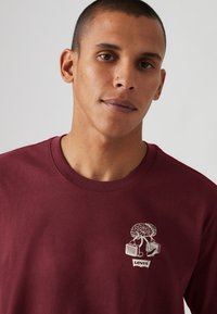 Bordeaux bomulds T-shirt med et hvidt grafisk logo på brystet, der viser et træ og sko, med et klassisk rundhalset design.