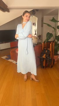 Γυναίκα με ανοιχτό μπλε μακρύ φόρεμα και μπεζ τακούνια στέκεται σε σαλόνι με ξύλινο πάτωμα, φυτά και διπλωμένες ξύλινες καρέκλες κοντά.
