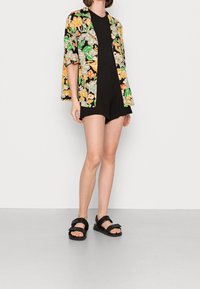 Mujer vestida con un mono negro y una camisa abierta con un patrón floral en tonos verdes, amarillos y naranjas, combinada con sandalias negras, de pie sobre un suelo gris.