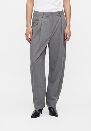 Persoon die een grijze hoge taille wijde broek met plooien draagt, zwarte puntige haklaarzen en een wit ingestopt topje.
