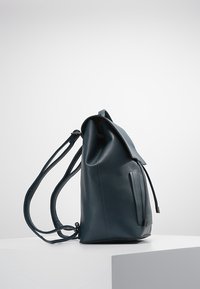 Ciemnozielony skórzany plecak o wyraźnej strukturze, z zamknięciem typu ściągacz oraz regulowanymi paskami. Boczna kieszeń z akcentem w postaci zamka błyskawicznego.