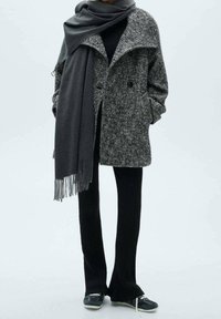 Manteau à motif pied-de-poule gris avec un col surdimensionné, associé à une écharpe gris foncé. Pantalon noir côtelé et chaussures foncées avec des semelles claires.