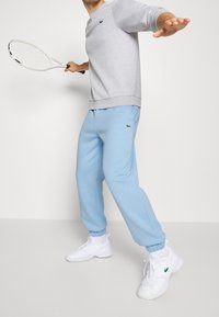 Ljusgrå sweatshirt med långa ärmar och grön logotyp, tillsammans med ljusblå sweatpants och vita träningsskor, håller en tennisracket.
