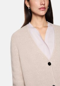 Hellbeige gestrickte Strickjacke mit lockerer Passform, V-Ausschnitt und schwarzem Knopfverschluss. Darunter ist eine weiche lilafarbene Bluse sichtbar.