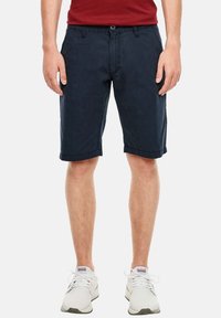 Muž v námořnických modrých kraťasech po kolena, bílých teniskách a červeném tričku, stojící na pozadí čistě bílé barvy.