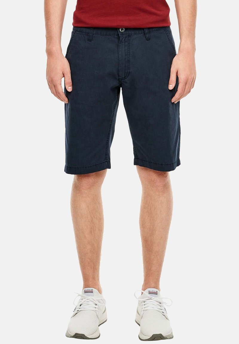 Muž v námořnických modrých kraťasech po kolena, bílých teniskách a červeném tričku, stojící na pozadí čistě bílé barvy.