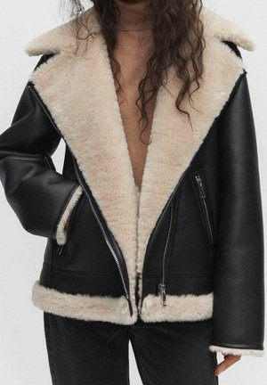 Personne portant une veste en cuir noire avec une doublure en peau lainée beige, des poches zippées et un col roulé beige en dessous.