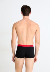 Schwarze Boxershorts mit rotem elastischem Bund und "HUGO"-Logo; glatte Textur und figurbetontes Design, von hinten gezeigt.