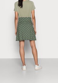 Žena stojící s viditelným zády, oblečená v zelené vzorované sukni, světle zeleném tričku s krátkým rukávem a bílých teniskách Reebok.