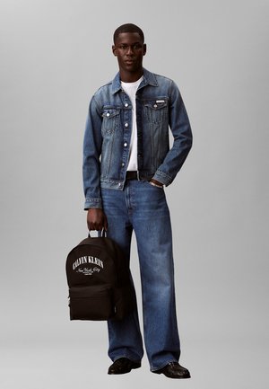 Giovane uomo con giacca e jeans di denim blu, t-shirt bianca, che tiene uno zaino nero Calvin Klein su uno sfondo grigio uniforme.