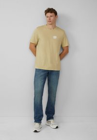 Beige katoenen t-shirt met korte mouwen en een klein wit logo op de borst, gedragen met blauwe denim jeans en lichte sneakers.