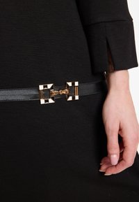 Nahaufnahme einer Hand, die neben einem schwarzen Gürtel mit einer geometrischen gold-weißen Schnalle auf einem schwarzen Kleidungsärmel mit Schlitzdetail ruht.