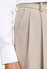 Pantaloni beige sartoriali con frontale plissettato e passanti per cintura, abbinati a una camicia bianca con polsini. Il tessuto presenta una texture liscia e una finitura ricercata.