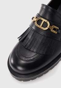 Black leather loafers featuring a textured finish, decorative fringing, and a gold chain accent on the strap. Thick black rubber sole.