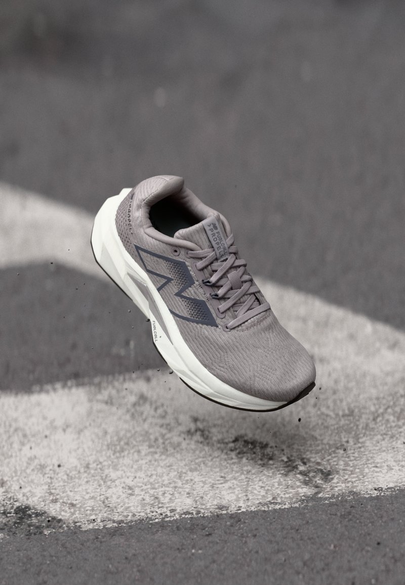 Γκρι αθλητικό παπούτσι για τρέξιμο με λευκή σόλα, στον αέρα πάνω από άσφαλτο και μια λευκή γραμμή ζωγραφισμένη, υπογραμμίζοντας την κίνηση και το ελαφρύ σχεδιασμό.