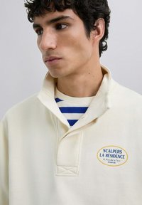 Suéter color crema con cuello, que presenta un logotipo bordado en azul y amarillo. Debajo, se puede ver una camisa a rayas azul y blanca.