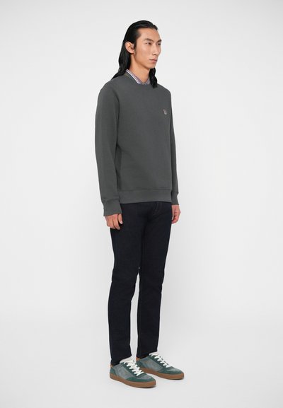 Vyras su ilgais juodais plaukais, vilkintis tamsiai pilką džemperį, tamsias džinsus, turkio ir žalios spalvos sportinius batelius, stovi priešais šviesiai baltą foną.