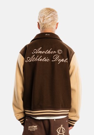 Persona con cabello rubio corto vistiendo una chaqueta universitaria marrón y beige con el texto "Another Athletic Dept." en la espalda y pantalones marrones a juego.
