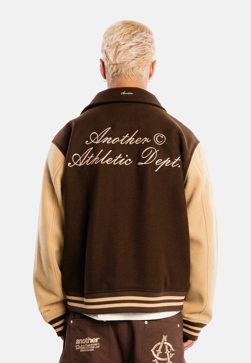 Personne aux cheveux blonds courts portant une veste universitaire marron et beige avec le texte "Another Athletic Dept." dans le dos et un pantalon marron assorti.