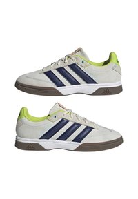 Paire de baskets Adidas basses en suède beige avec des rayures bleu marine, une doublure intérieure vert citron, une semelle marron et une semelle intermédiaire blanche, vues de côté.